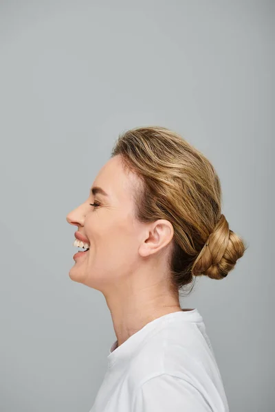 Радісна приваблива жінка зі зібраним світлим волоссям позує на сірому фоні і щиро сміється — стокове фото