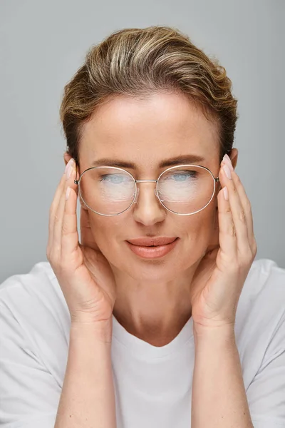 Attraktive blonde Frau in lässiger Kleidung mit gesammelten Haaren posiert mit ihrer Brille vor grauem Hintergrund — Stockfoto