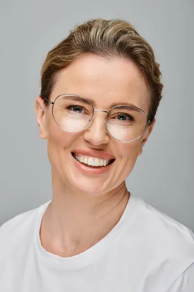 Joyful beautiful woman with blonde collected hair and glasses smiling at camera on gray backdrop — Stock Photo