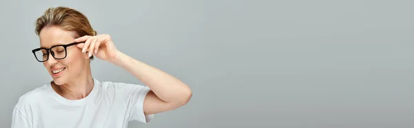 Mulher atraente alegre com cabelo loiro e óculos posando em pano de fundo cinza e olhando para longe, banner — Fotografia de Stock