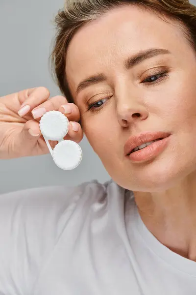 Gut aussehende fröhliche Frau mit blonden Haaren, die Kontaktlinsen hält und vor grauem Hintergrund wegschaut — Stockfoto