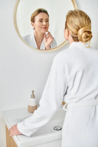 Alegre mulher bonita com cabelo loiro coletado em roupão de banho olhando para o espelho em seu banheiro — Fotografia de Stock