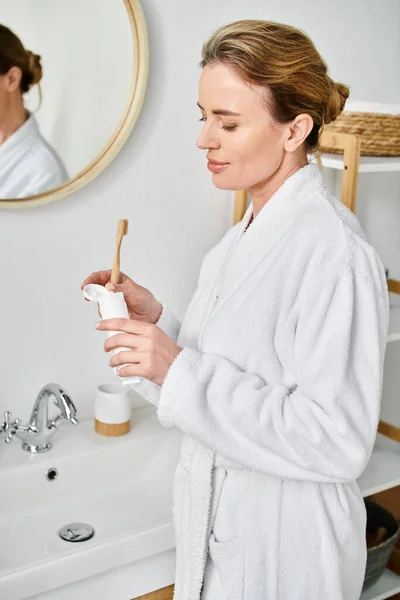 Mulher loira atraente com cabelo coletado em roupão de banho escovando os dentes na frente do espelho — Fotografia de Stock