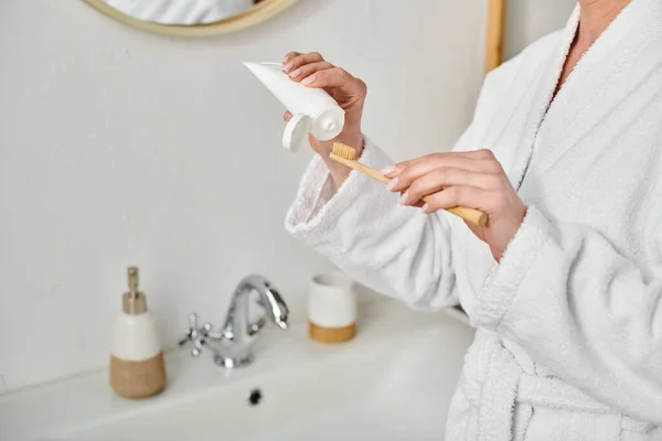 Vue recadrée de la femme adulte en peignoir confortable mettre de la pâte dentaire sur sa brosse près du miroir — Photo de stock