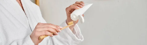 Cropped view of adult woman in comfy bathrobe putting tooth paste on her brush near mirror, banner — Stock Photo
