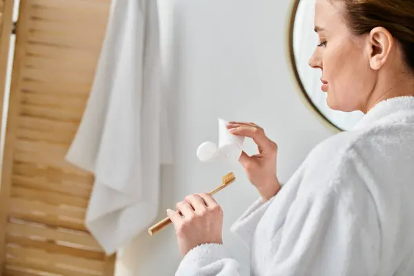 Добре виглядає блондинка в халаті чистить зуби перед дзеркалом у ванній — стокове фото
