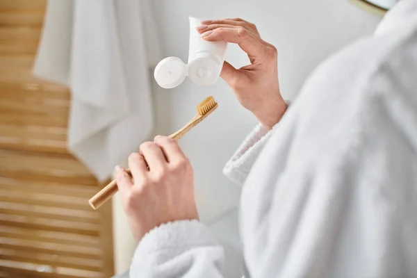 Vista recortada de mujer adulta en albornoz acogedor poniendo pasta de dientes en su cepillo cerca del espejo - foto de stock