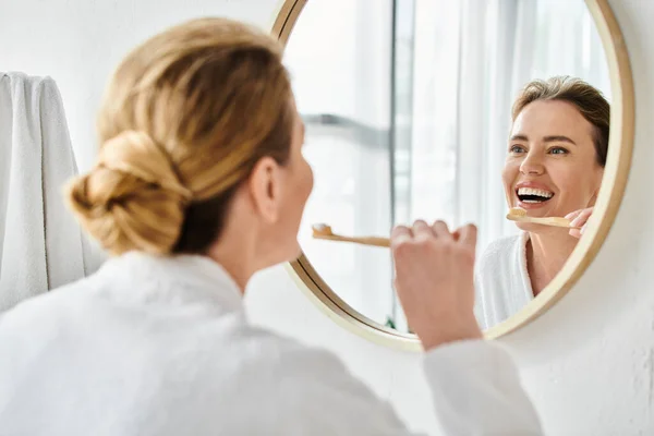 Attractive joyous woman with blonde hair in bathrobe brushing her teeth and looking at mirror — Stock Photo