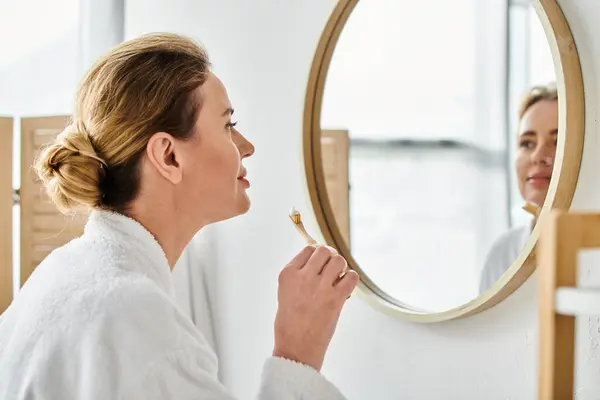 Mulher loira bonita em roupão de banho escovando os dentes na frente do espelho em seu banheiro — Fotografia de Stock