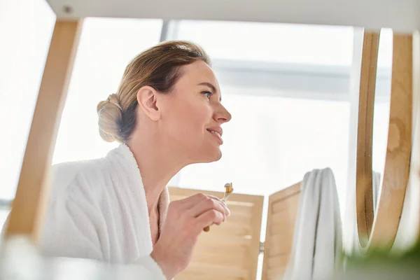 Appealing joyful woman with blonde hair in bathrobe brushing her teeth and looking at mirror — Stock Photo