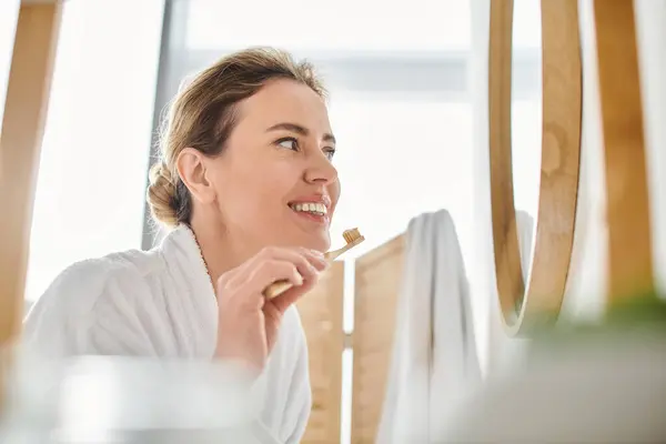 Attraktive, fröhliche Frau mit blonden Haaren im Bademantel, die sich die Zähne putzt und in den Spiegel schaut — Stockfoto