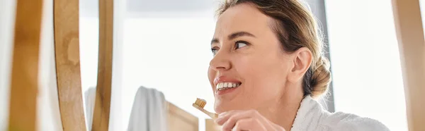 Приваблива радісна жінка з світлим волоссям в халаті чистить зуби і дивиться на дзеркало, банер — стокове фото