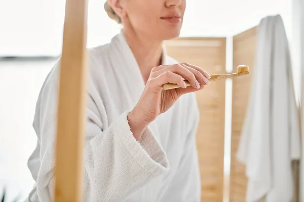 Vue recadrée de la femme adulte en peignoir confortable mettre de la pâte dentaire sur sa brosse près du miroir — Photo de stock