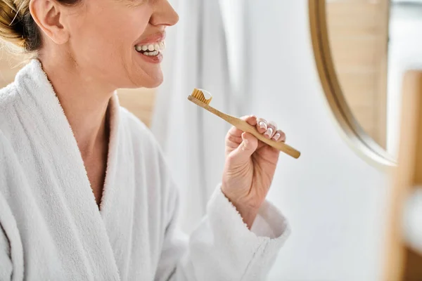 Vista recortada de la alegre mujer adulta en albornoz poniendo pasta de dientes en su cepillo cerca del espejo - foto de stock