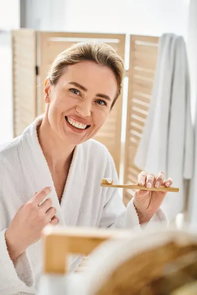 Séduisante femme gaie en peignoir avec des cheveux blonds regardant la caméra tout en se brossant les dents — Photo de stock