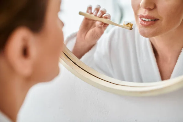 Vue recadrée de la femme adulte en peignoir blanc mettre de la pâte dentaire sur sa brosse près du miroir — Photo de stock