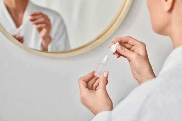 Cropped view of adult woman in bathrobe using her eye drops in front of mirror in bathroom — Stock Photo