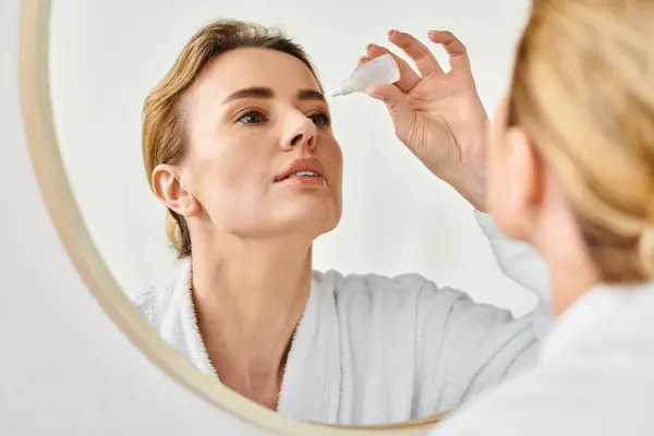 Beautiful blonde woman with collected hair in bathrobe putting in eye drops in her bathroom — Stock Photo