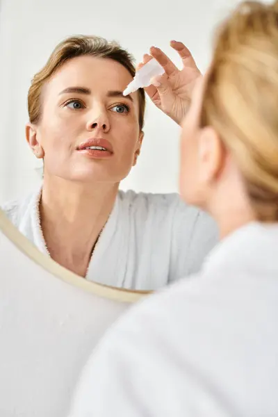 Schöne blonde Frau mit gesammelten Haaren im Bademantel und Augentropfen in ihrem Badezimmer — Stockfoto