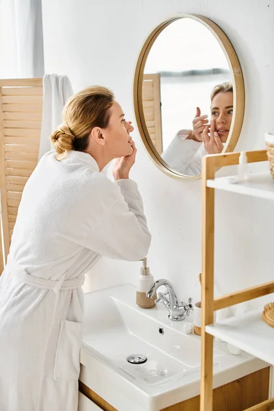 Attractive woman in white comfy bathrobe wearing her contact lenses near mirror in bathroom — Stock Photo