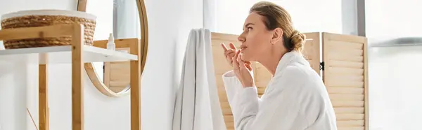 Привлекательная женщина в белом удобном халате носить ее контактные линзы возле зеркала в ванной комнате, баннер — стоковое фото
