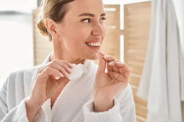 Cheerful appealing woman in white bathrobe cleaning her teeth with dental floss in bathroom — Stock Photo
