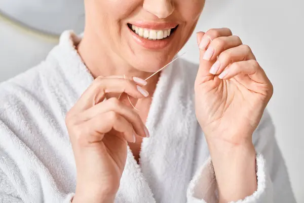 Cropped view of jolly adult woman in bathrobe cleaning her teeth with dental floss in bathroom — Stock Photo