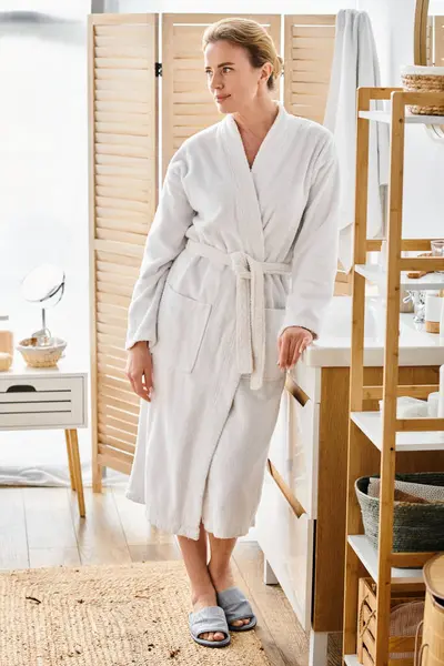 Atractiva mujer alegre con el pelo rubio en albornoz blanco posando en su baño y mirando hacia otro lado - foto de stock