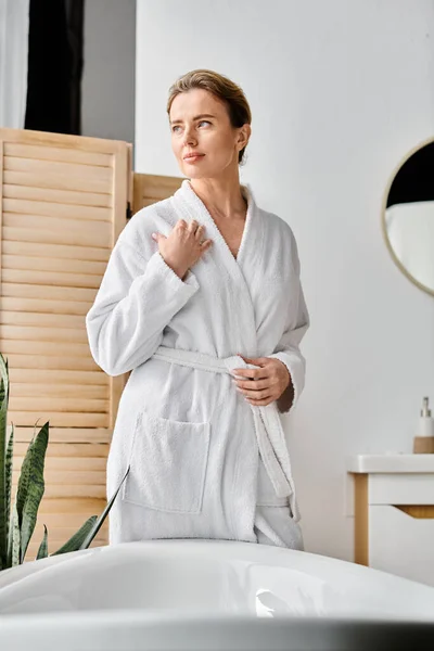 Гарна весела жінка з світлим волоссям у білому затишному халаті позує біля своєї ванни — стокове фото