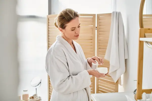 Appealing blonde woman with collected hair in bathrobe holding face cream in her hands in bathroom — Stock Photo