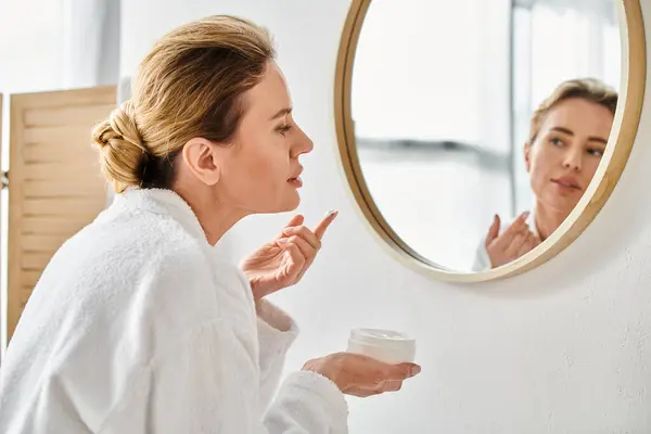 Belle femme blonde en peignoir blanc confortable appliquant crème pour le visage tout en salle de bain à la maison — Photo de stock