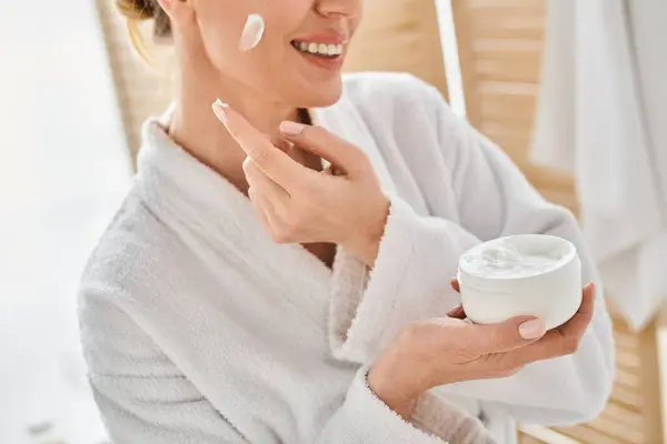 Cropped view of cheerful woman in comfy bathrobe applying her face cream in bathroom at her home — Stock Photo