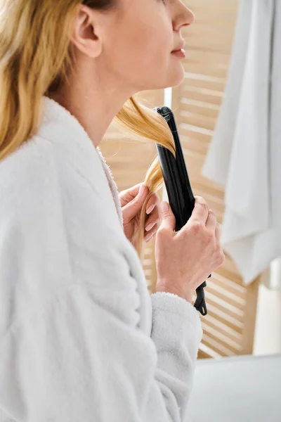 Vista recortada de mujer rubia en cómodo albornoz usando plancha plana en su cabello en el baño - foto de stock