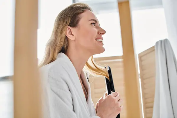 Cheerful appealing blonde woman in cozy bathrobe using flat iron on her hair while in bathroom — Stock Photo