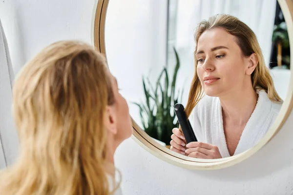Atractiva mujer rubia en acogedor albornoz usando plancha plana en su cabello y mirando al espejo - foto de stock