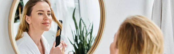 Cheerful appealing blonde woman in cozy bathrobe using flat iron on her hair while in bathroom — Stock Photo
