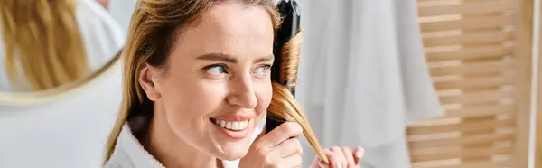 Mujer atractiva alegre en albornoz acogedor usando plancha plana en su cabello mientras está en el baño, pancarta - foto de stock