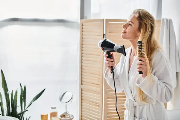 Appealing blonde joyous woman in bathrobe using hair dryer and brush on her long hair at home — Stock Photo