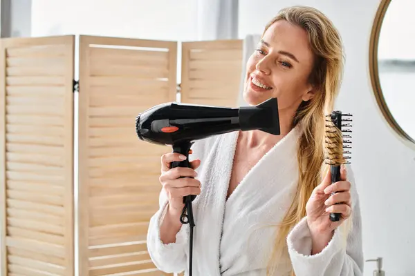 Attraente gioiosa donna bionda in accappatoio utilizzando asciugacapelli e spazzolare i capelli lunghi a casa — Foto stock