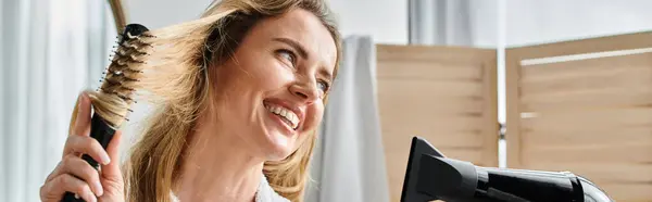 Attractive joyous woman in bathrobe using hair dryer and brush on her long hair at home, banner — Stock Photo