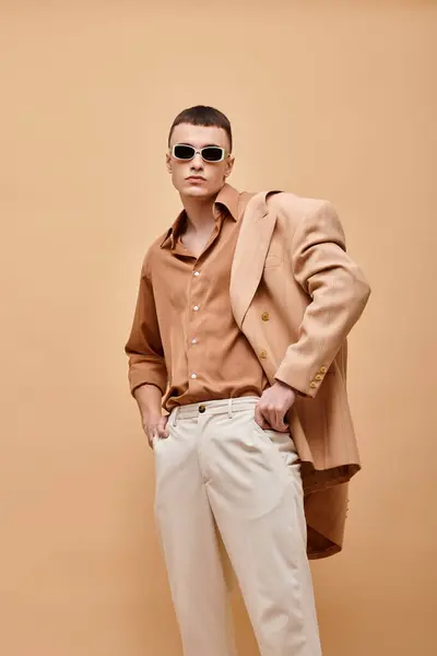Hombre de moda en chaqueta beige en hombro, camisa, pantalones y gafas de sol posando sobre fondo beige - foto de stock
