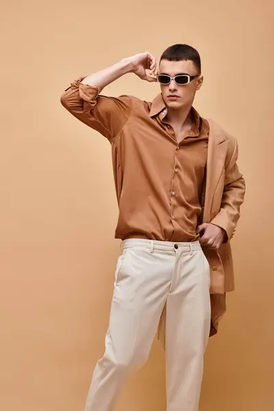Hombre con clase en chaqueta beige en hombro, camisa, pantalones y gafas de sol posando sobre fondo beige - foto de stock