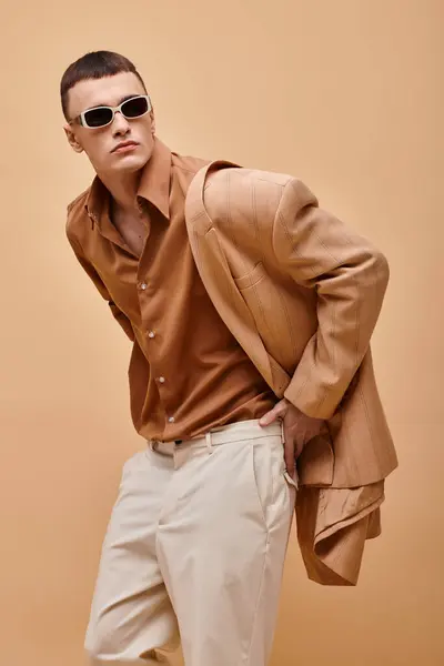 Elegant man in beige jacket on shoulder, shirt, pants and sunglasses posing on beige background — Stock Photo