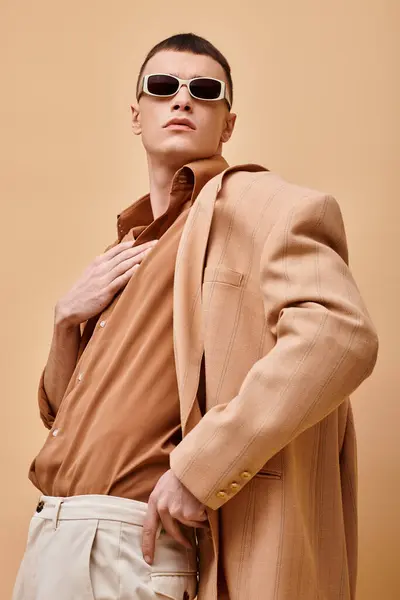 Portrait of man in beige jacket and shirt with sunglasses and hand near collar on beige backdrop — Stock Photo