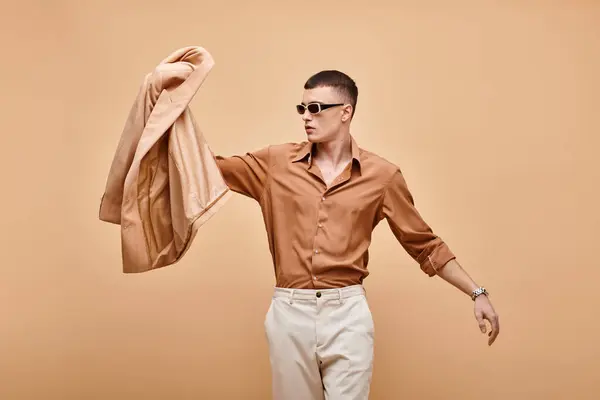 Modeaufnahme eines Mannes in beigem Hemd mit Sonnenbrille und Jacke in der Hand vor beigem Hintergrund — Stockfoto