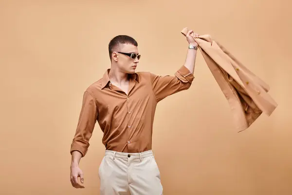 Fashion shot of man in beige shirt with sunglasses and throwing jacket away on beige backdrop — Stock Photo