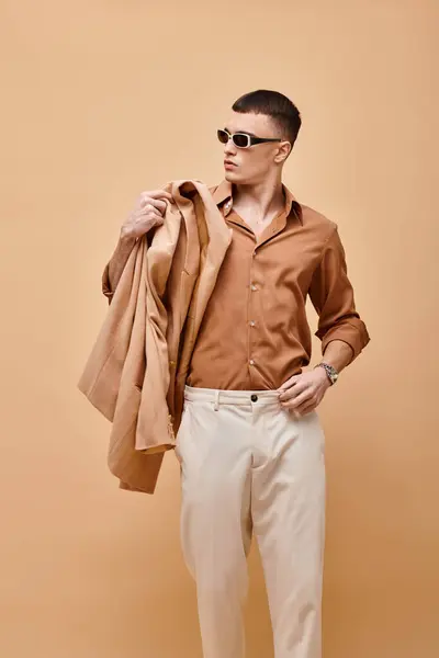 Fashion photo of man in beige shirt with sunglasses and holding jacket in hand on beige backdrop — Foto stock
