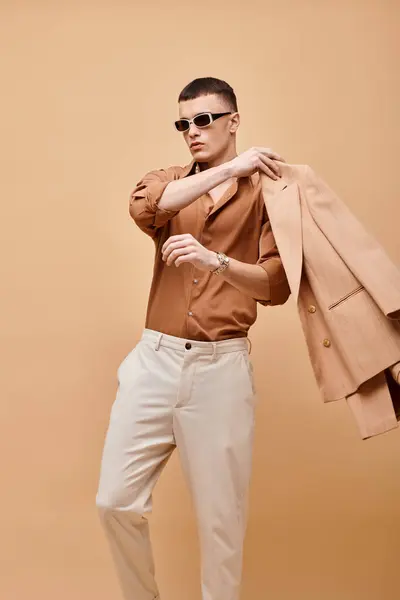 Portrait of man in beige shirt with sunglasses and putting his jacket on  beige backdrop — Stock Photo