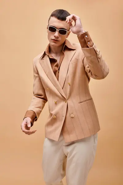 Selbstbewusster Mann in beiger Jacke, Hemd, Hose und Sonnenbrille posiert auf beigem Hintergrund — Stockfoto