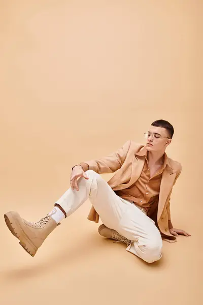 Stylish man in 20s in beige jacket and glasses sitting on peachy beige background — Stock Photo